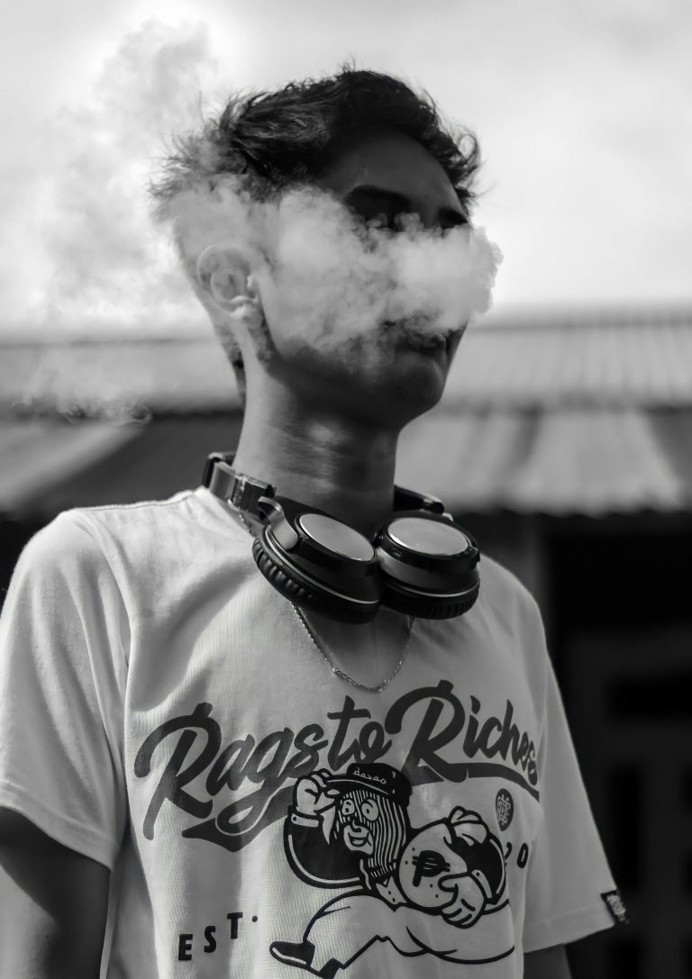 A young man has headphones around his neck, vapes and exhales a cloud, looking edgy and stylish.