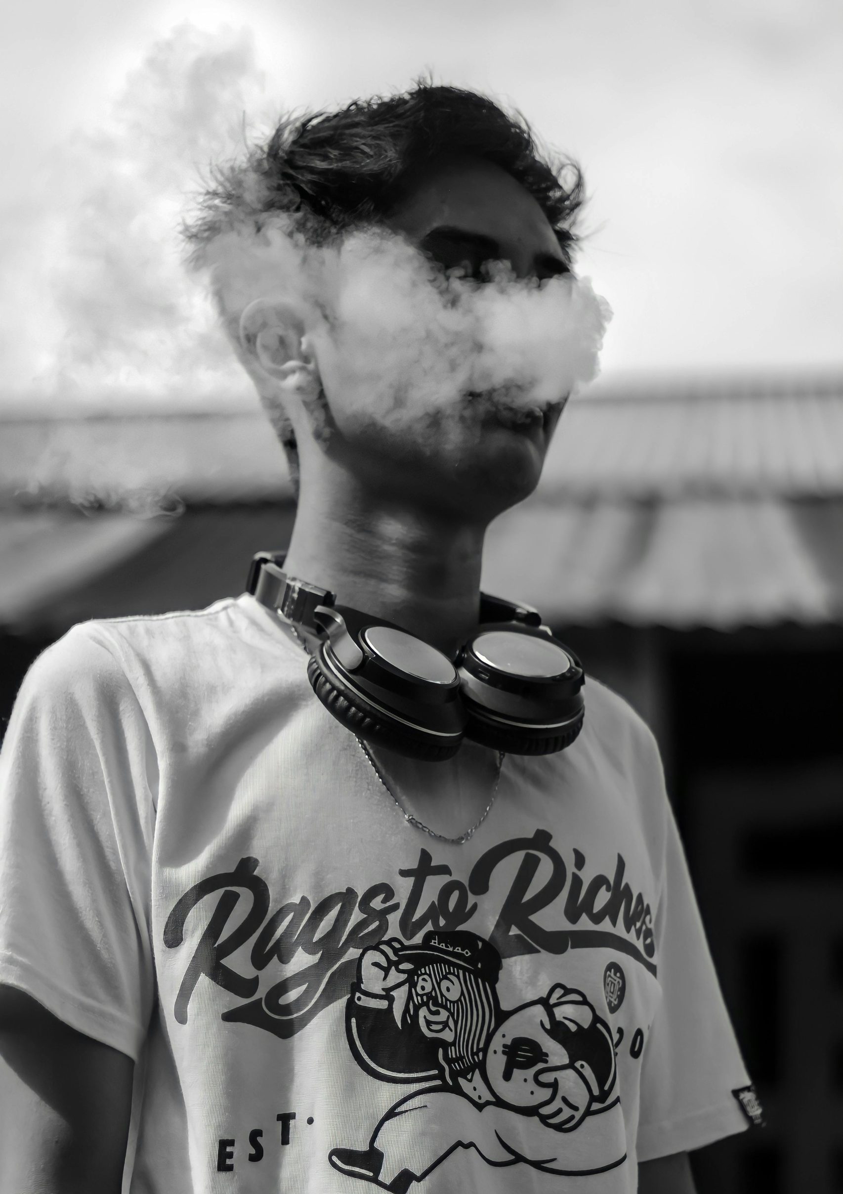 A young man has headphones around his neck, vapes and exhales a cloud, looking edgy and stylish.
