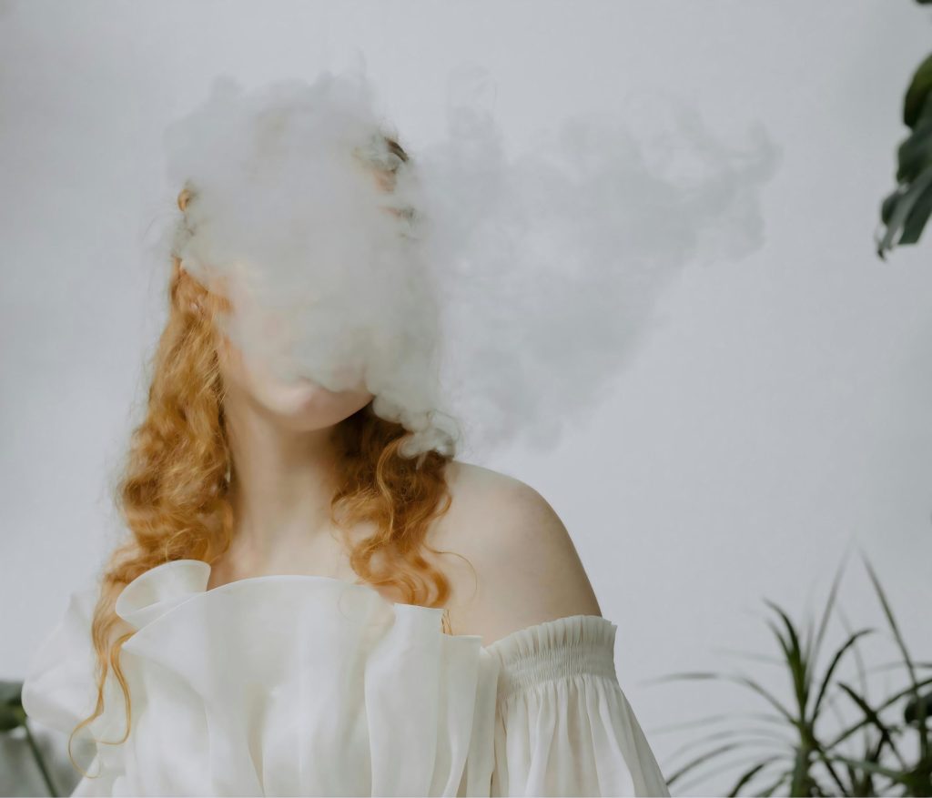 Vape cloud chasing photo: Red-haired person exhaling thick white vapor (cloud chasing effect) with green plants behind.