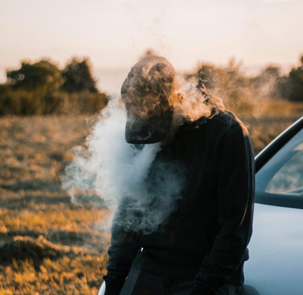 A person releases dense vape clouds outdoors at dusk, with grassland and a car in the background.